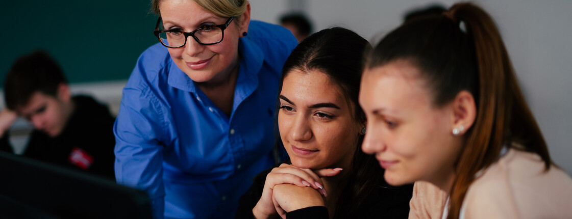 Eine Lehrerin zeigt zwei Schüler*innen etwas am Computer-Bildschirm.