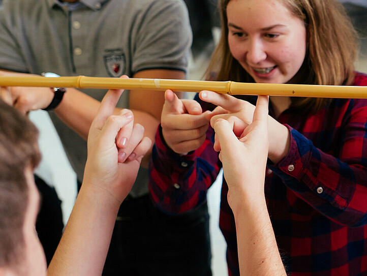 Schüler*innen halten gemeinsam, jede*r mit zwei Fingern, einen Stab in der Hand und balancieren diesen.