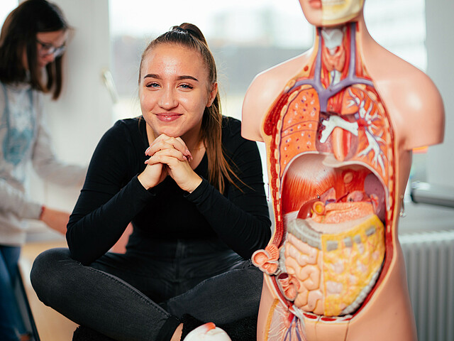 Eine Schülerin sitzt neben einem anatomischen Korpus des menschlichen Körpers und lächelt in die Kamera.