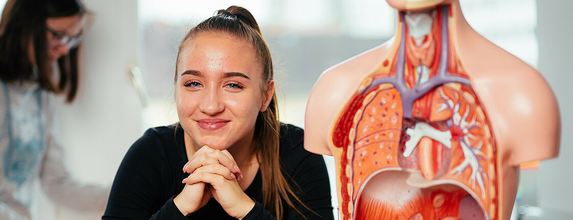 Eine Schülerin sitzt neben einem anatomischen Korpus des menschlichen Körpers und lächelt in die Kamera.