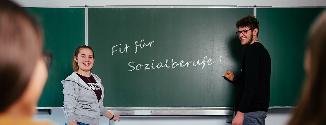 Eine Schülerin und ein Schüler stehen an der Tafel auf der "Fit für Sozialberufe" steht.