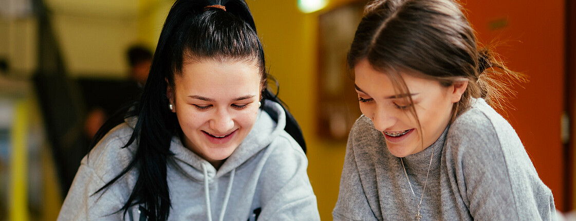 Zwei Schülerinnen sitzen gemeinsam vor einem Textbuch, lernen und lächeln dabei.